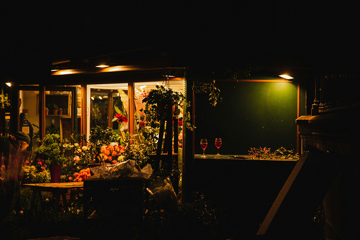Ambiente nocturno en una floristería iluminada, con copas de vino sobre una barra rodeada de plantas y flores variadas