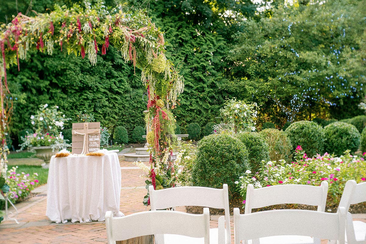 Ceremonia de boda en jardín con arco floral decorado, sillas blancas y set de bienvenida